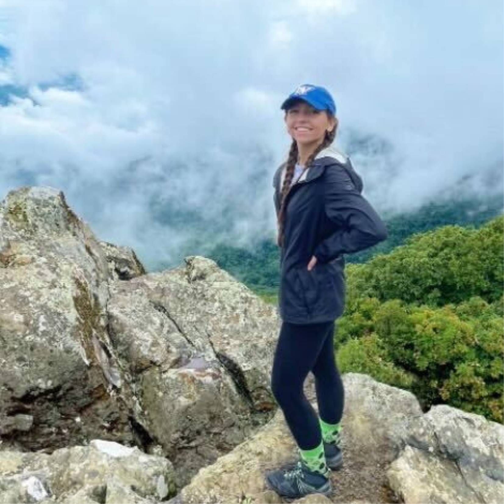 Image of female standing on top of large rocks overlooking a cloudy, tree filled valley.